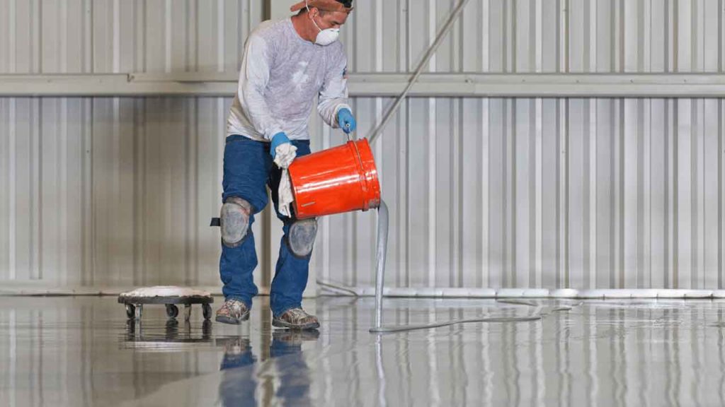 Tradesperson in protective gear pouring epoxy resin from a bucket onto a warehouse floor