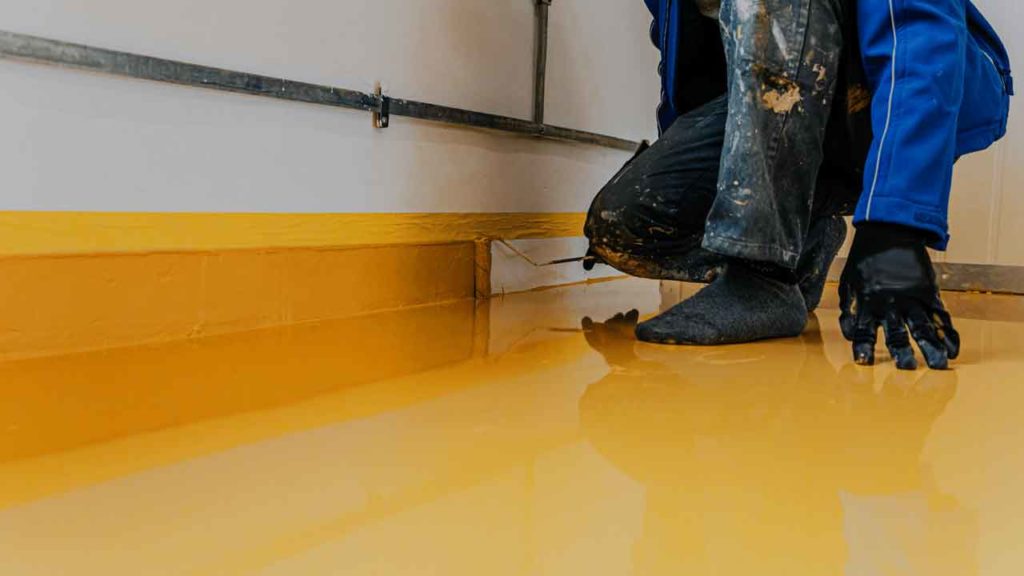 Tradesperson kneeling to smooth yellow epoxy coating along a wall edge with a trowel